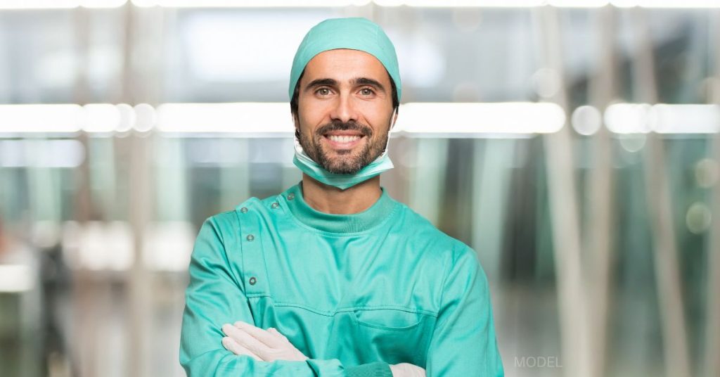 A male doctor is standing with his arms crossed smiling and wearing a pair of mint green scrubs and gloves. (model)