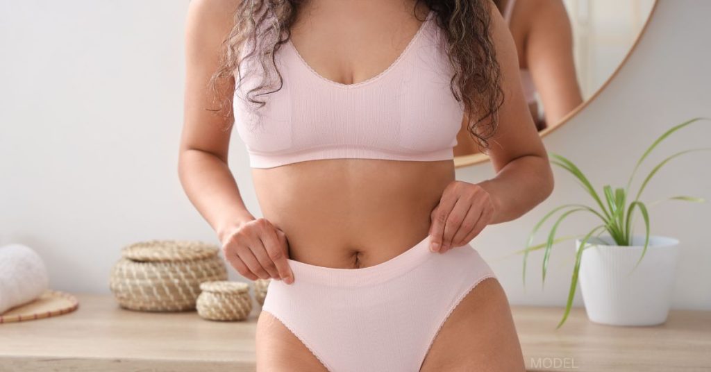 A woman with brown hair is wearing a pink bra and underwear with her hands against her hips. (model)